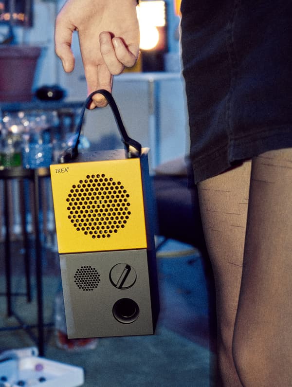 Una mujer con medias negras y minifalda sujetando un altavoz portátil IKEA FREKVENS de color amarillo y negro.
