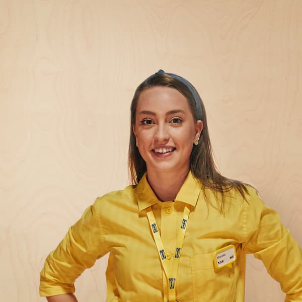 Una mujer de cabello castaño sonriendo con la camiseta amarilla, el colgante y la etiqueta con el nombre de compañera de IKEA.
