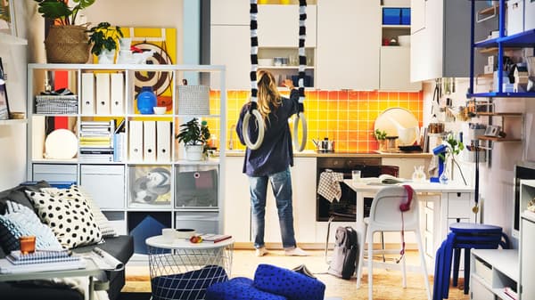 Una mujer en su cocina abriendo un gabinete, se observan estantes, mesa y gabinetes blancos. También se ve un sofa color gris con cojines decorativos encima de este.
