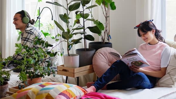 Una mujer en una cama apilable UTÅKER leyendo un libro. Detrás de una combinación de plantas y cortinas que divide la habitación, se encuentra un hombre.