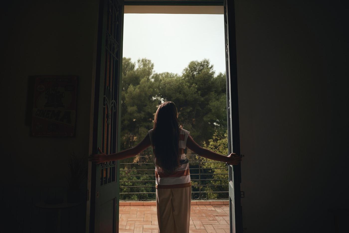 Una mujer mirando de frente a la salida de una puerta, donde ve árboles y un piso de tierra.