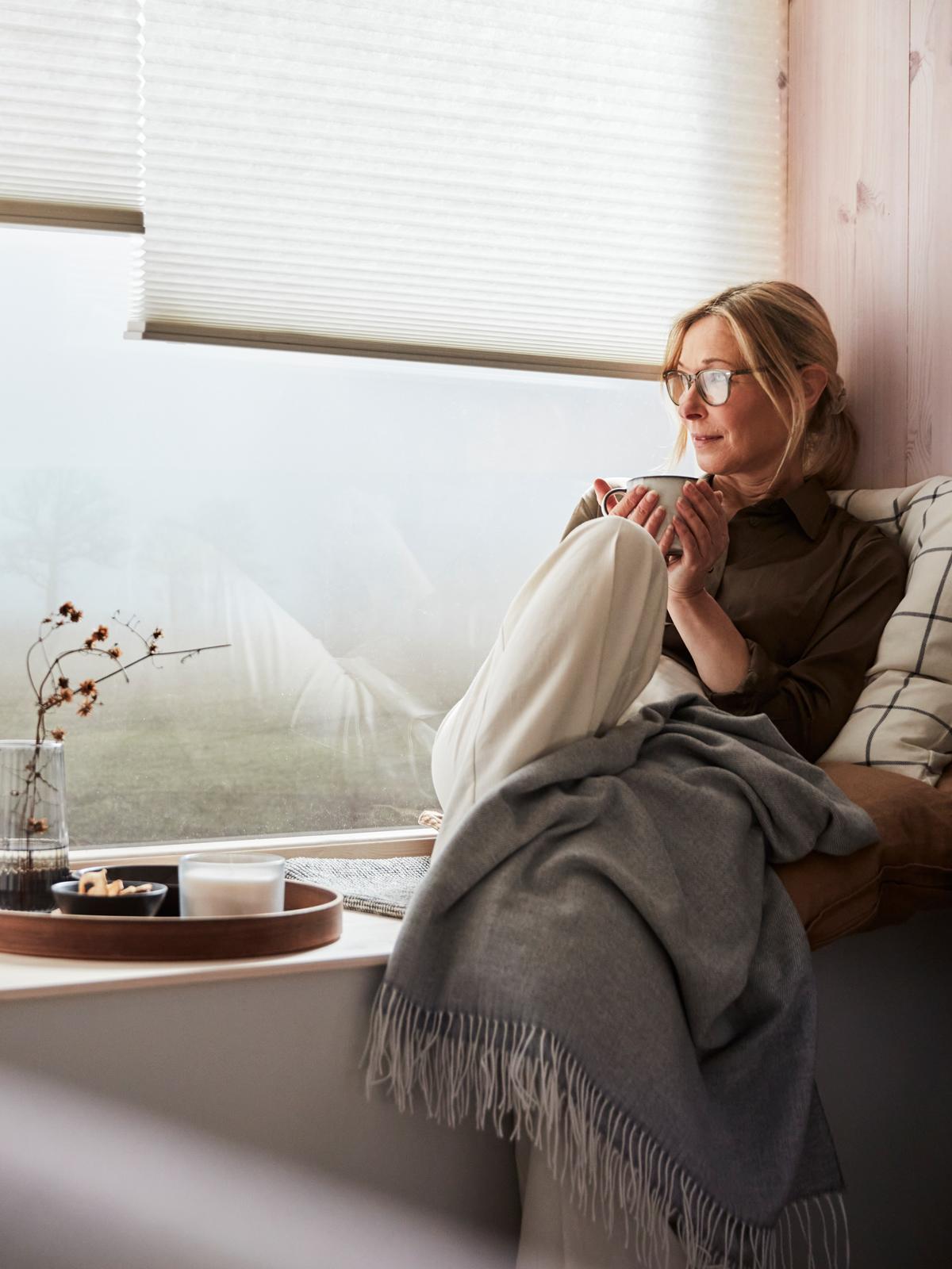 Una mujer se sienta en el alféizar de una amplia ventana y mira un día brumoso, mientras se cubre con una manta, un cojín y una taza de té.
