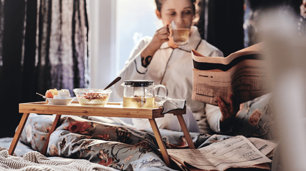Una mujer sentada en una cama con una banjeda con alimentos y café para tomar desayuno en cama.