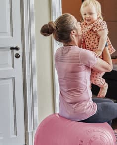 Una mujer sentada en una pelota de ejercicio rosa, sosteniendo y sonriendo a un bebé risueño que lleva un pijama con estampados de corazones.