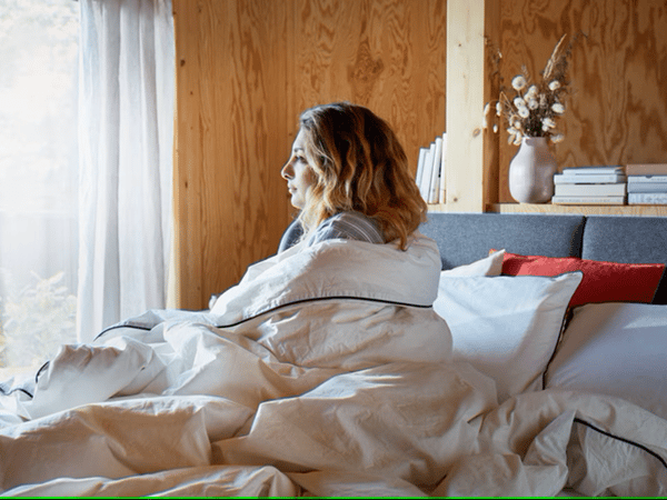 Una mujer sentada sobre una cama envuelta en un edredón blanco.