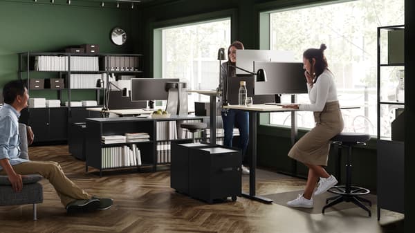 Una mujer sentada trabajando en un computador portátil sobre un escritorio blanco en una sala vacía, rodeada de cajas de embalaje IKEA sin desarmar. Al fondo se ve un colgador de ropa y un panel con papeles. 