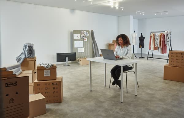 Una mujer sentada trabajando en un computador portátil sobre un escritorio blanco en una sala vacía, rodeada de cajas de embalaje IKEA sin desarmar. Al fondo se ve un colgador de ropa y un panel con papeles. 