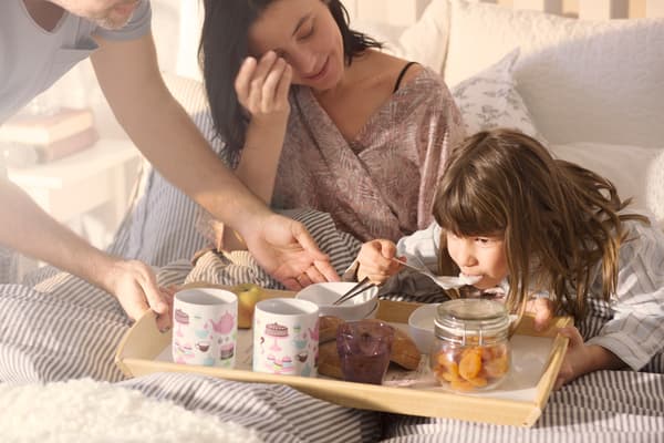 Una mujer y su hija tomando desayuno en la cama, con una bandeja. Comen cosas con cucharas y hay dos tazas.