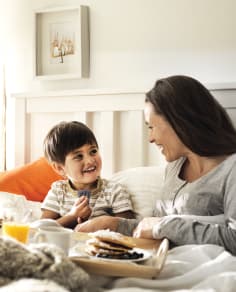 Una mujer y un niño pequeño sonriendo y mirándose en la cama, con una bandeja de desayuno con panqueques y jugo de naranja entre ellos. 