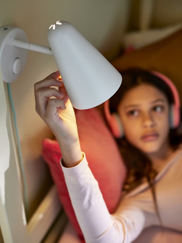Una niña con audifonos rosas reclinado contra un cojín rosa ajusta una lámpara de pared FUBBLA LED blanca sobre ella.