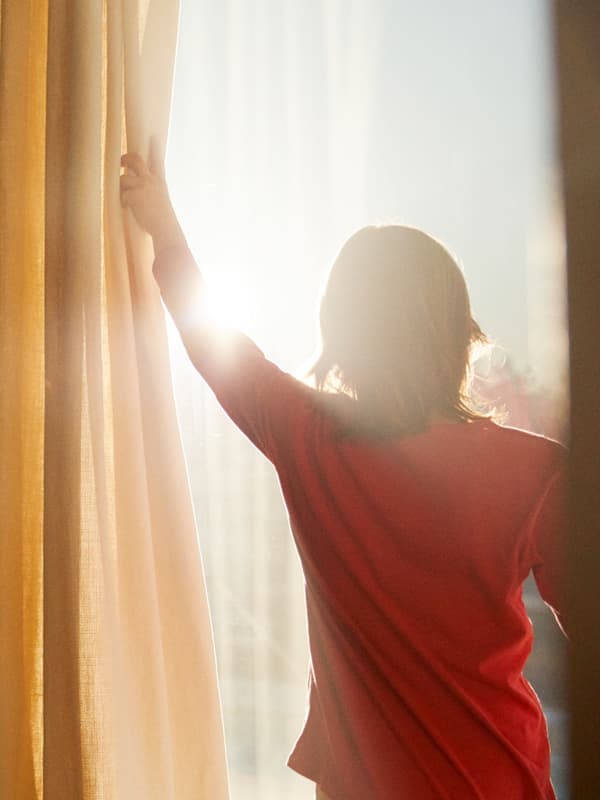 Una niña con una camiseta roja corre las cortinas amarillas y blancas para mostrar el sol que entra por la ventana.