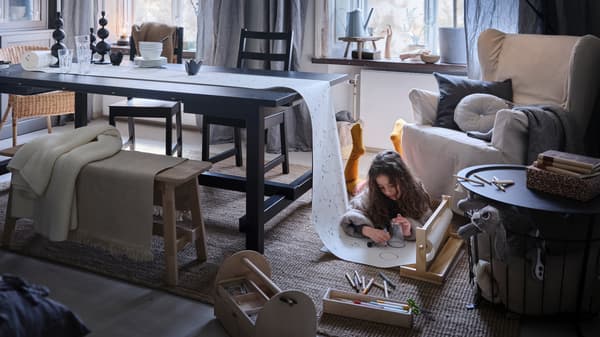 Una niña en el suelo con una alfombra dibujando en un rollo de papel, con un extremo sobre una mesa extensible negra NORDVIKEN .