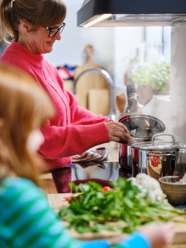 Una niña está junto a una barra de cocina mientras una adulta con un jersey rojo remueve una olla IKEA 365+ de acero inoxidable en la parte posterior de una estufa de inducción.