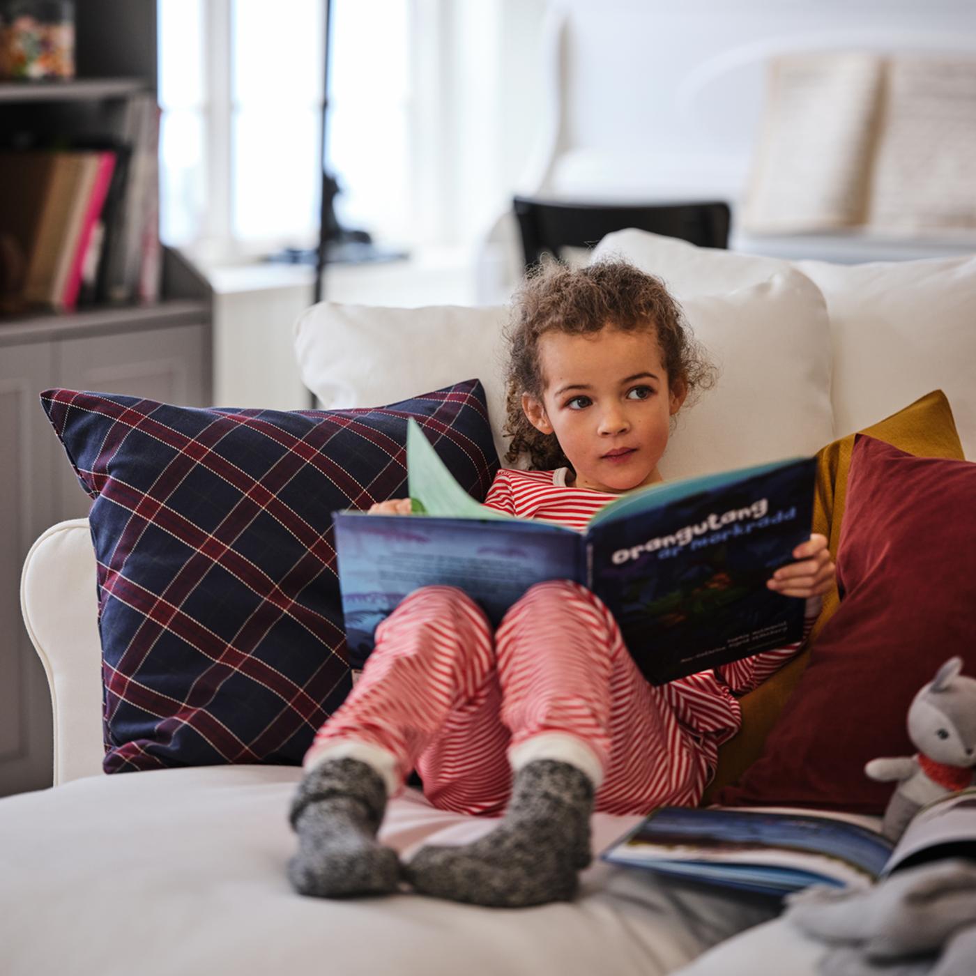 Una niña pequeña en pijama sentada cómodamente con un libro en un sofá blanco de cuatro plazas GRÖNLID con una chaise longue.