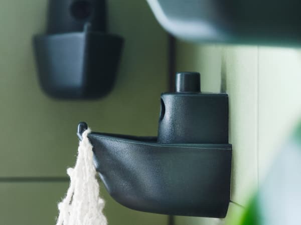 Una pared de azulejos verdes con tres ganchos verdes de KNOPARE que parecen un barco de juguete, uno de los cuales sostiene una bolsa de macramé blanca.