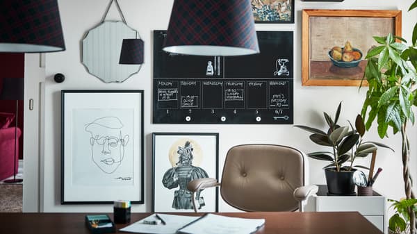 Una pared de una oficina doméstica cubierta con diferentes obras de arte. Hay una lámpara de techo sobre un escritorio y una silla.