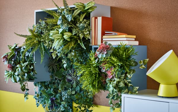Una parete decorata con numerose piante artificiali contenute in mobili e sopra diversi libri. Accanto, una lampada gialla.