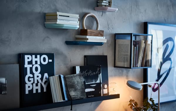 Una parete effetto cemento con una minivetrina BARKHYTTAN contenente libri. Accanto, una mensola MALMBÄCK con libri aperti e chiusi e una clessidra. 