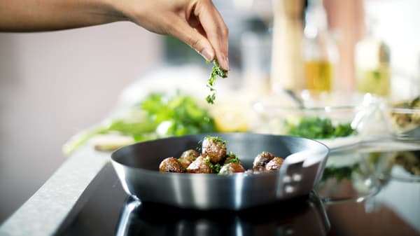Una persona aliñando con hierbas las albóndigas HUVUDROLL que se cocinan en un sartén metálico sobre una encimera eléctrica. 
