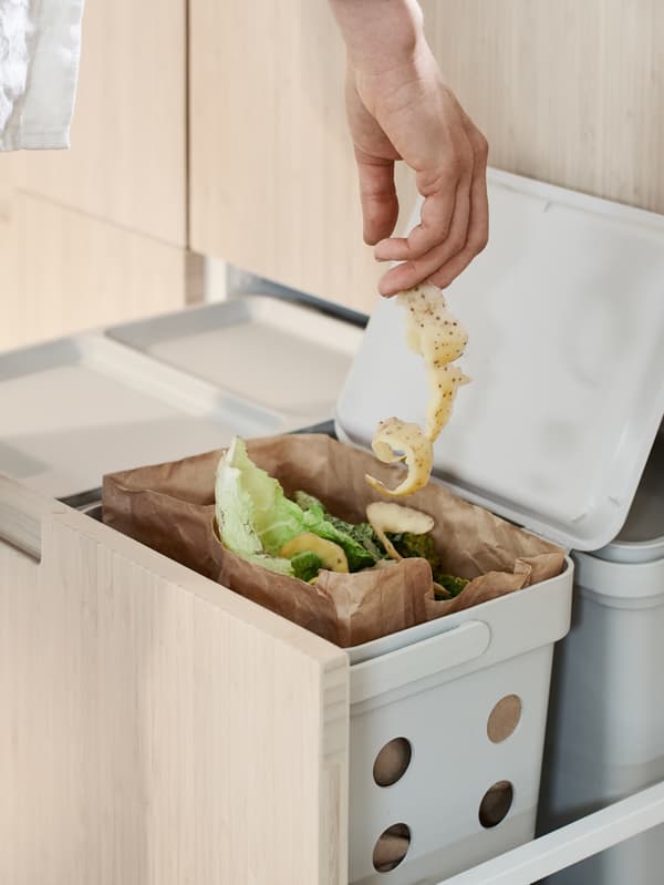 Una persona añade una cáscara de verdura a una bolsa de papel marrón para residuos de alimentos en un contenedor de clasificación de residuos HÅLLBAR dentro de un cajón alto MAXIMERA.