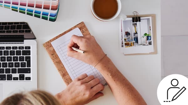 Una persona annota qualcosa sul taccuino sulla sua scrivania. Accanto a lei un PC, una tazza e una scala di colori pantone - IKEA