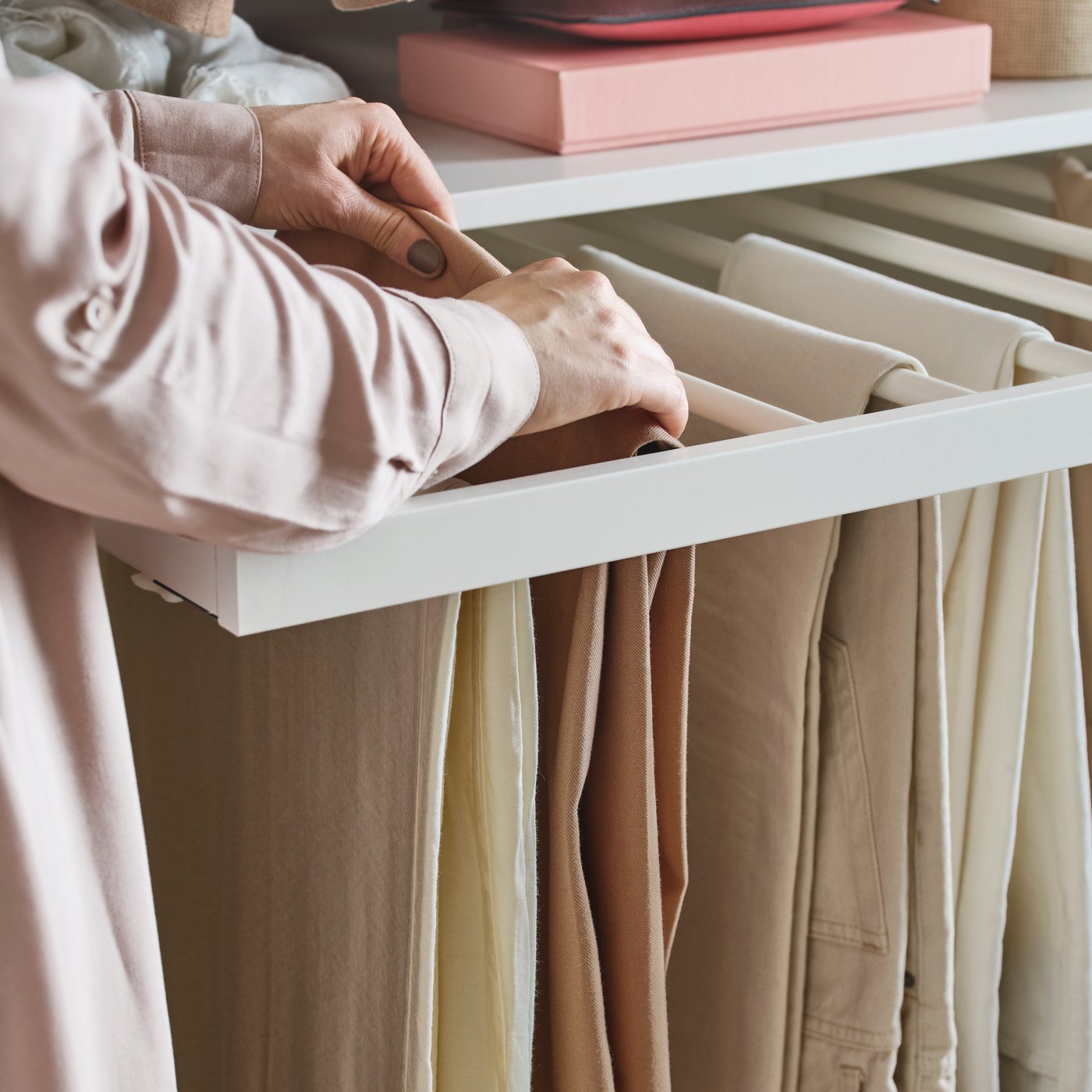 Una persona che indossa una camicia rosa appende un paio di pantaloni beige su un portapantaloni estraibile KOMPLEMENT già pieno di pantaloni.