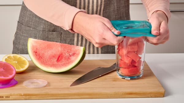 Una persona coloca una campana de alimentos de silicona ÖVERMÄTT en turquesa sobre un vaso con sandía troceada que está sobre una tabla de cortar.