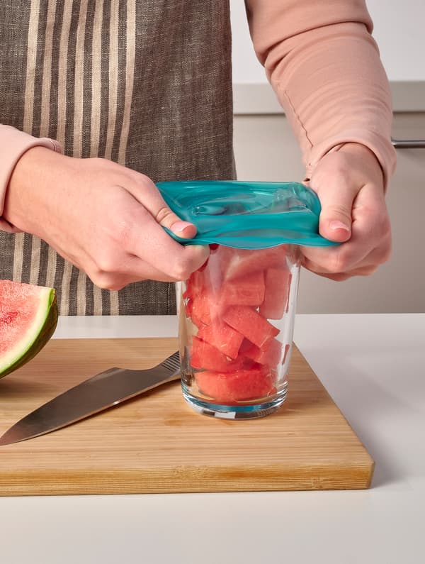 Una persona coloca una cubierta para alimentos ÖVERMÄTT de silicona turquesa sobre un vaso de sandía picada que se encuentra sobre una tabla de cortar.
