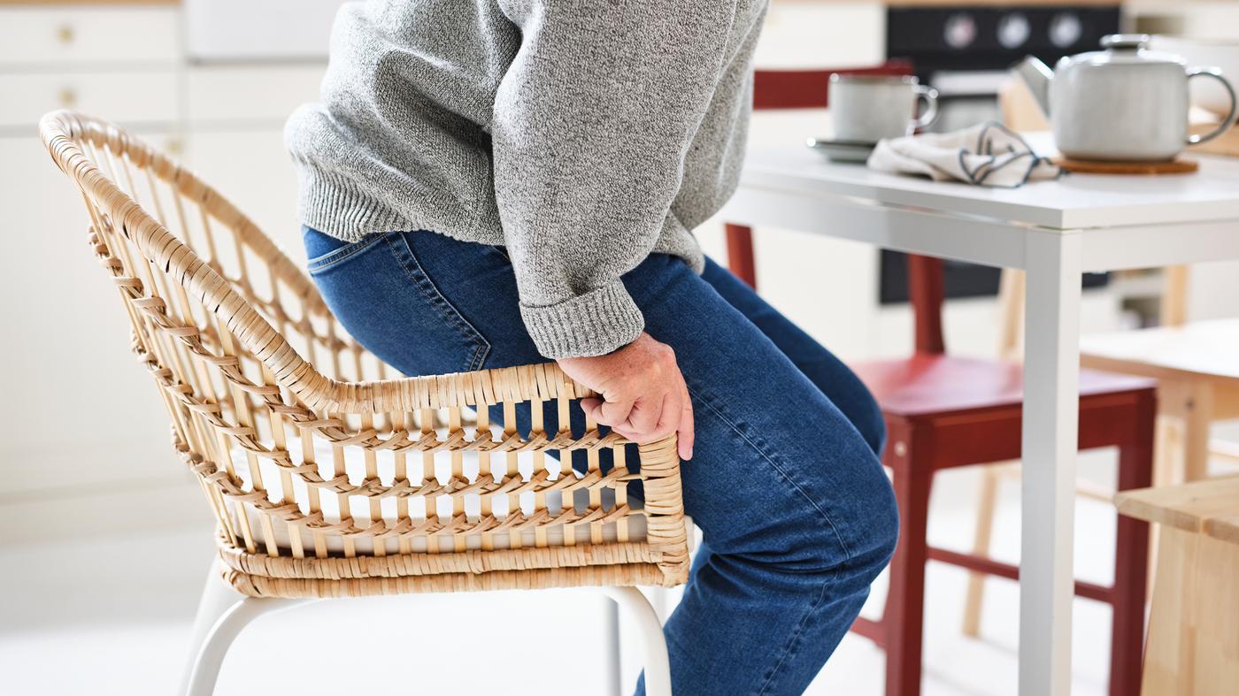 Una persona con jeans azules y un jersey gris se levanta de una silla de ratán con un cojín.