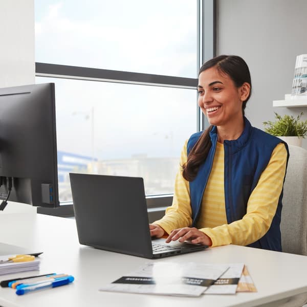 Una persona con una camisa de compañero/a de IKEA se sienta en un escritorio blanco trabajando en la computadora detrás de la ventana.