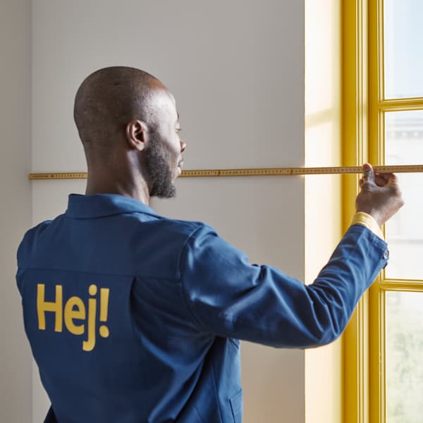 Una persona con una chaqueta azul midiendo una sección de una pared blanca con un metro plegable junto a una ventana amarilla.