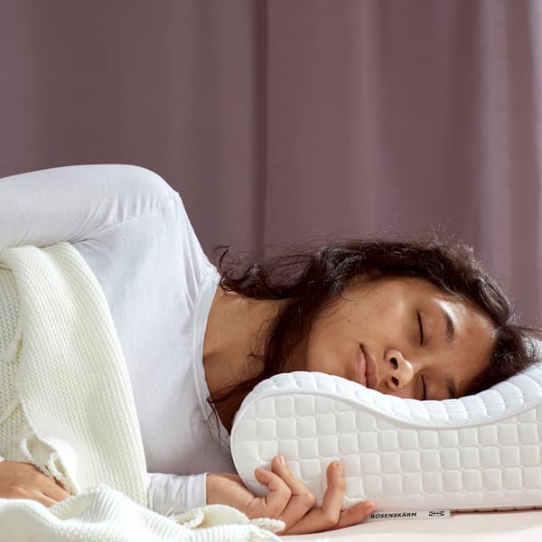 Una persona con una maglietta bianca dorme su un cuscino ergonomico ROSENSKÄRM su un letto dietro il quale c'è una tenda lilla.