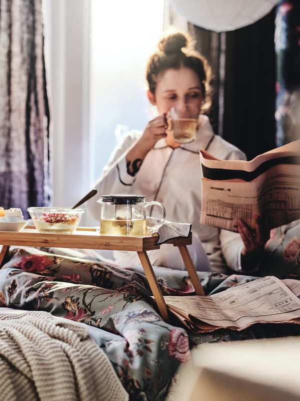 Una persona está desayunando en una cama cubierta con una funda nórdica de flores de color gris verdoso claro.
