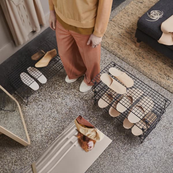 Una persona in piedi tra due rastrelliere per scarpe GREJIG in filo d’acciaio con sopra forme per scarpe. Sul pavimento e a lato ci sono due specchi.