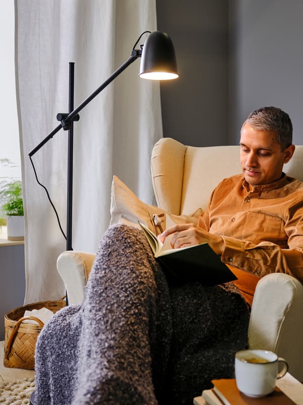 Una persona lee en un sillón orejero STRANDMON de Risane de color natural bajo una lámpara de pie/lectura SKURUP negra.
