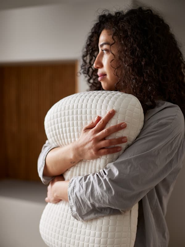 Una persona mira desde la distancia mientras abraza una almohada ergonómica KLUBBSPORRE en su pecho. La luz brilla en su rostro.