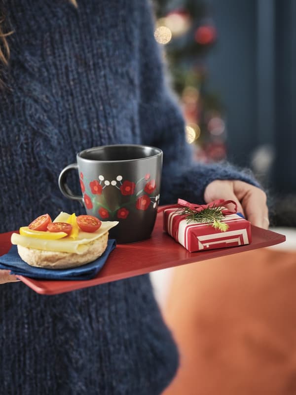 Una persona porta su un vassoio un piccolo pacchetto regalo, una tazza VINTERFINT e un dolcetto con frutta. Sullo sfondo, si intravede un albero di Natale.