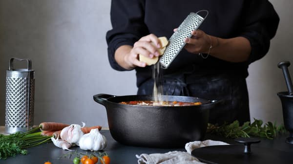 Una persona ralla queso sobre una cacerola VARDAGEN negra mientras prepara una sopa.