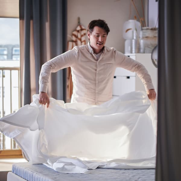 Una persona stende un lenzuolo su un materasso a molle VADSÖ rigido/azzurro, sopra un letto imbottito SLATTUM.