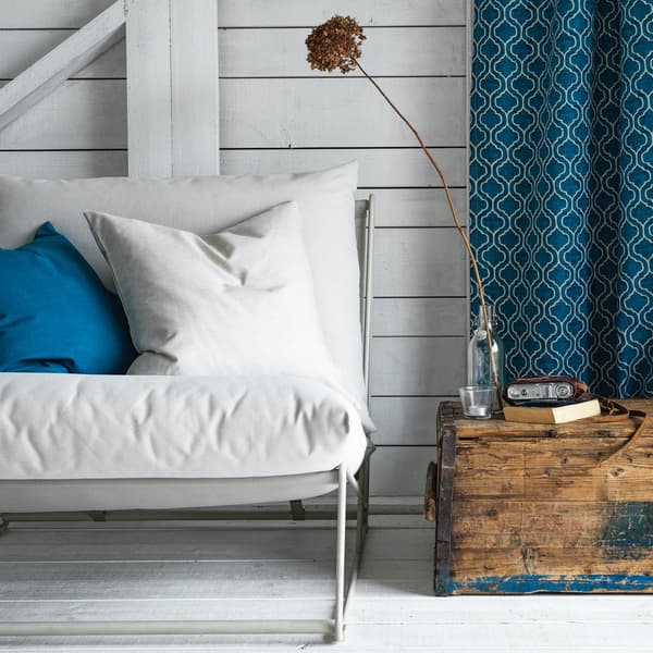 Una poltrona bianca con un cuscino bianco e uno blu. A lato, un baule in legno con sopra libri e un vaso con fiore.
