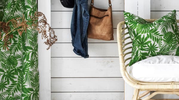 Una poltrona in rattan BUSKBO con un cuscino verde tropicale. A lato, una borsa e un indumento appeso.