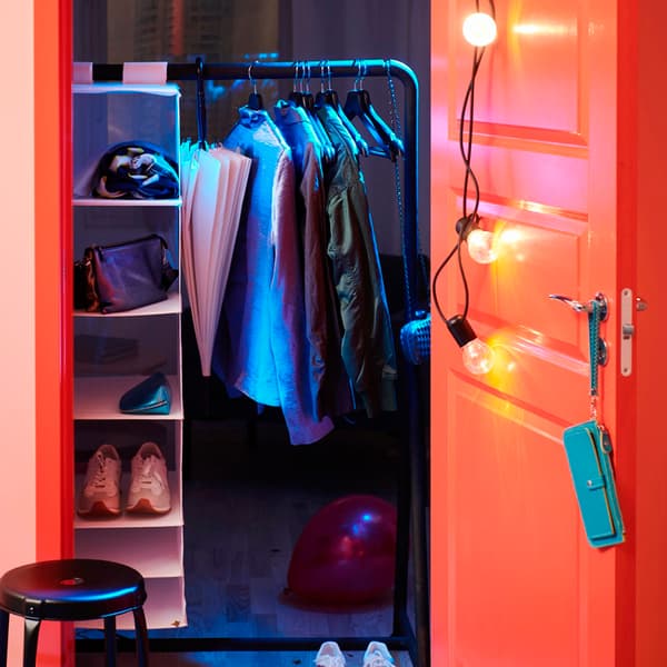 Una porta aperta colorata da luci che mostra uno stand appendiabiti nero con indumenti ordinati e un portatutto STUK. A lato, uno sgabello nero.