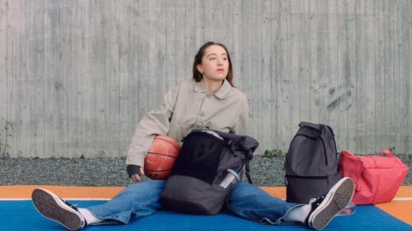 Una ragazza seduta a terra su un campo da basket, con in mano un pallone da basket, accanto agli zaini DRÖMSÄCK, VÄRLDENS e STARTTID.