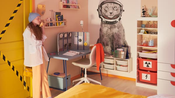 Una ragazza vicino alla porta di una stanza dai colori pastello con una scrivania LAGKAPTEN/TILLSLAG blu, una sedia da ufficio LÅNGFJÄLL, mobili e un letto - IKEA