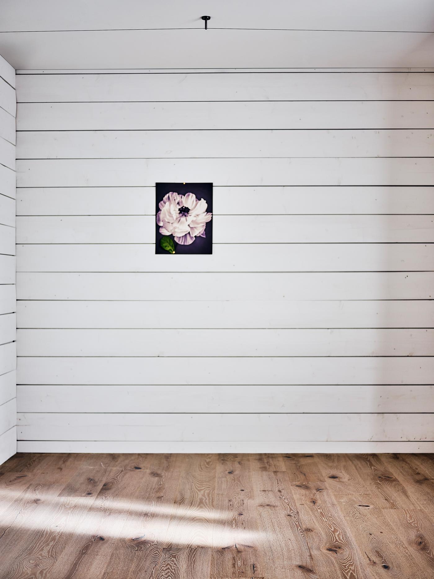 Una recámara vacía con paredes de paneles de madera blanca y piso de madera clara y un cuadro de una flor en la pared.