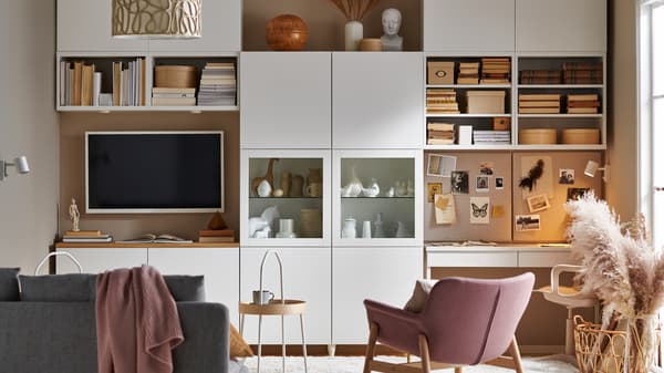 Una sala con un sistema de almacenamiento BESTÅ blanco que cubre una pared con almacenamiento abierto y oculto, un sofá, una televisión y una estación de trabajo.