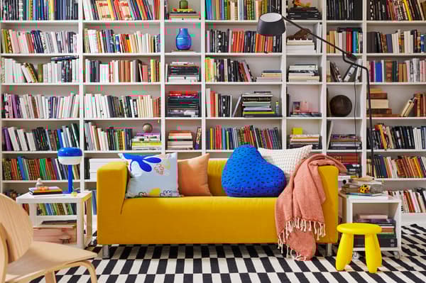 Una sala de estar con un sofá amarillo con cojines naranjos y azules. Frente a muebles de biblioteca lleno de libros.