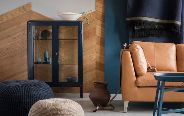 Una sala d’estar blava i marró amb un sofà, un reposapeus i una vitrina amb portes de vidre IKEA FABRIKÖR de color blau.
