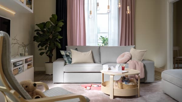 Una sala en tonos pastel con un sofá KIVIK gris claro, cortinas KALKFLY rosa claro y una mesa de centro BORGEBY.