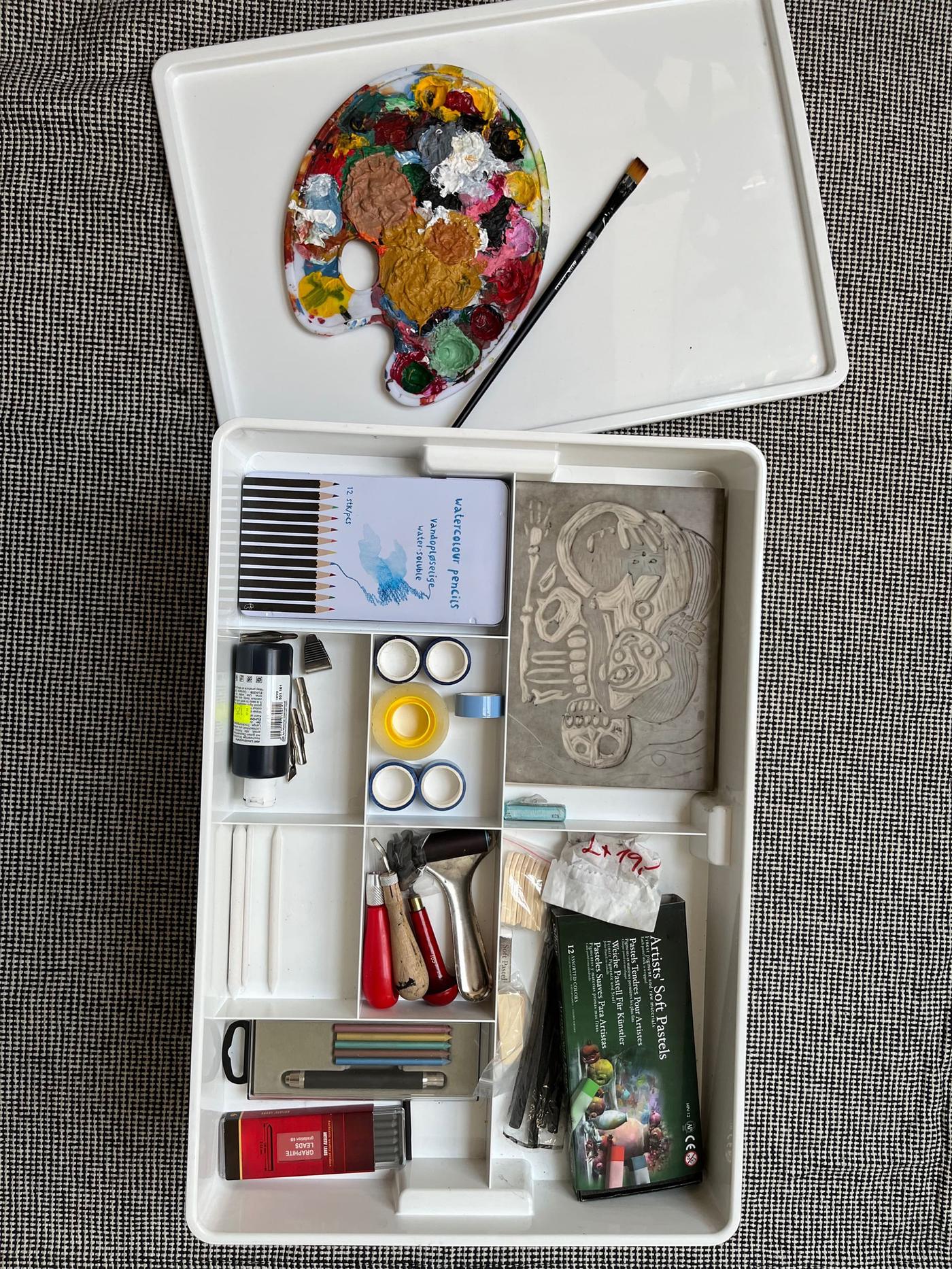 Una scatola contenitore KUGGIS di IKEA di colore bianco, aperta e vista dall'alto e al suo interno una serie di colori, tubetti di colore, matite, pennelli e il necessario per dipingere.